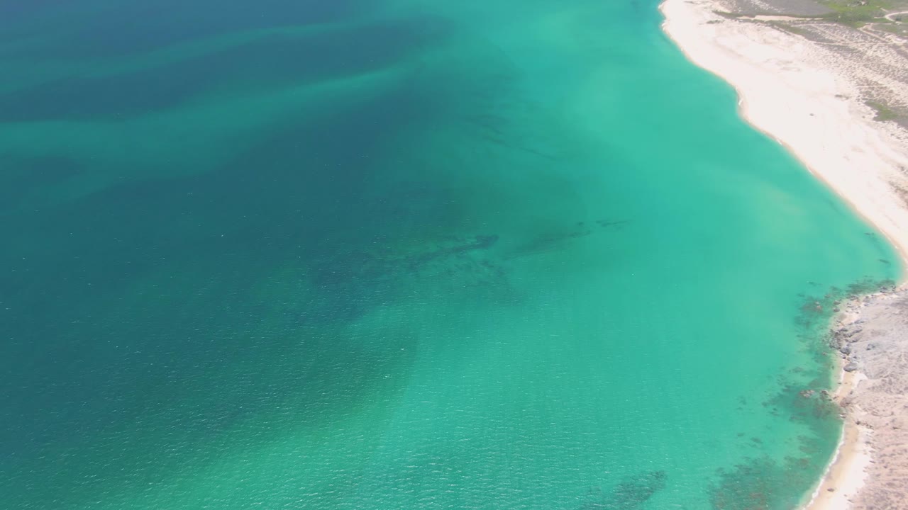 At 0:01, the Playa Miramar shoreline remains at the right edge while clear turquoise water reveals darker seabed patches and no visible people.