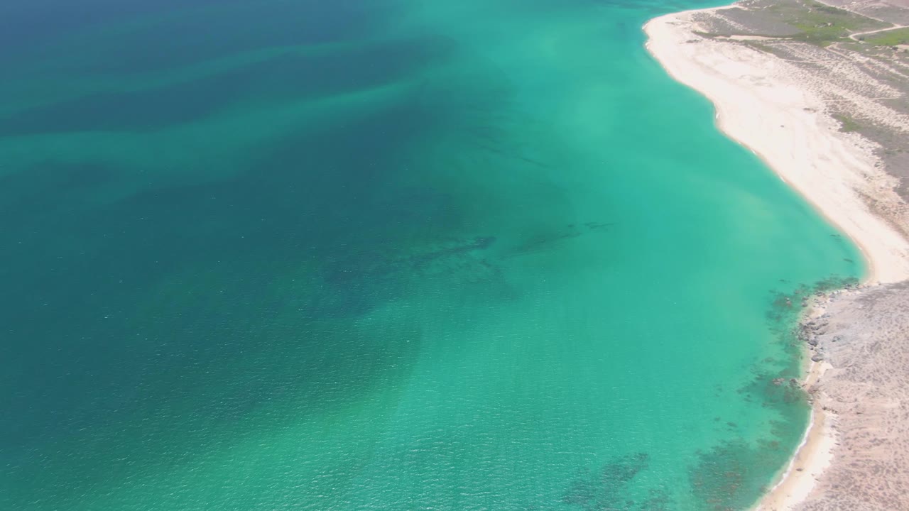 At 0:02 over Playa Miramar Los Cabos, the pale shoreline curves along the right side beside turquoise shallows and darker offshore water, with no visible people.