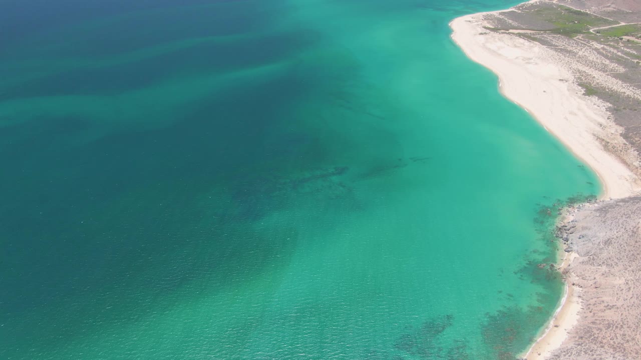 At 0:03, the aerial view shows calm blue-green water beside the sandy and rocky Playa Miramar coast, with faint seabed texture and no visible people.