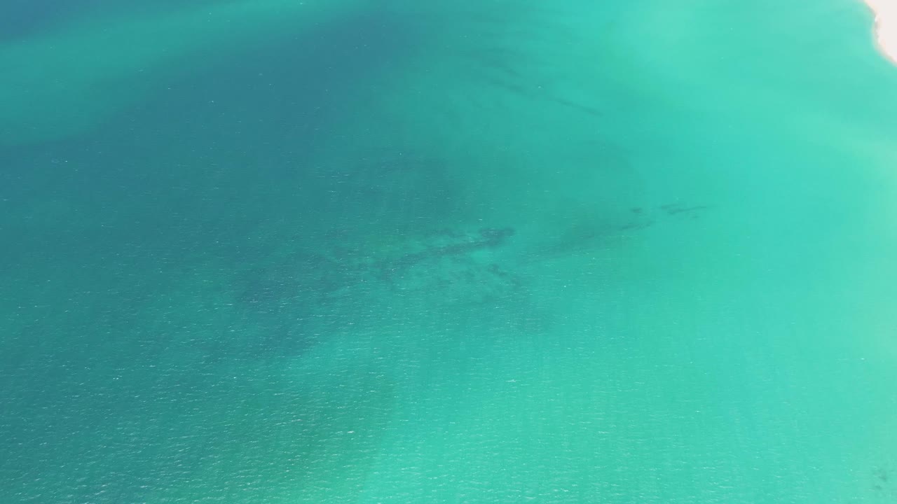 Clear turquoise shallows at Playa Miramar Los Cabos show a sandy bottom and soft darker areas beneath the surface, with no visible people.