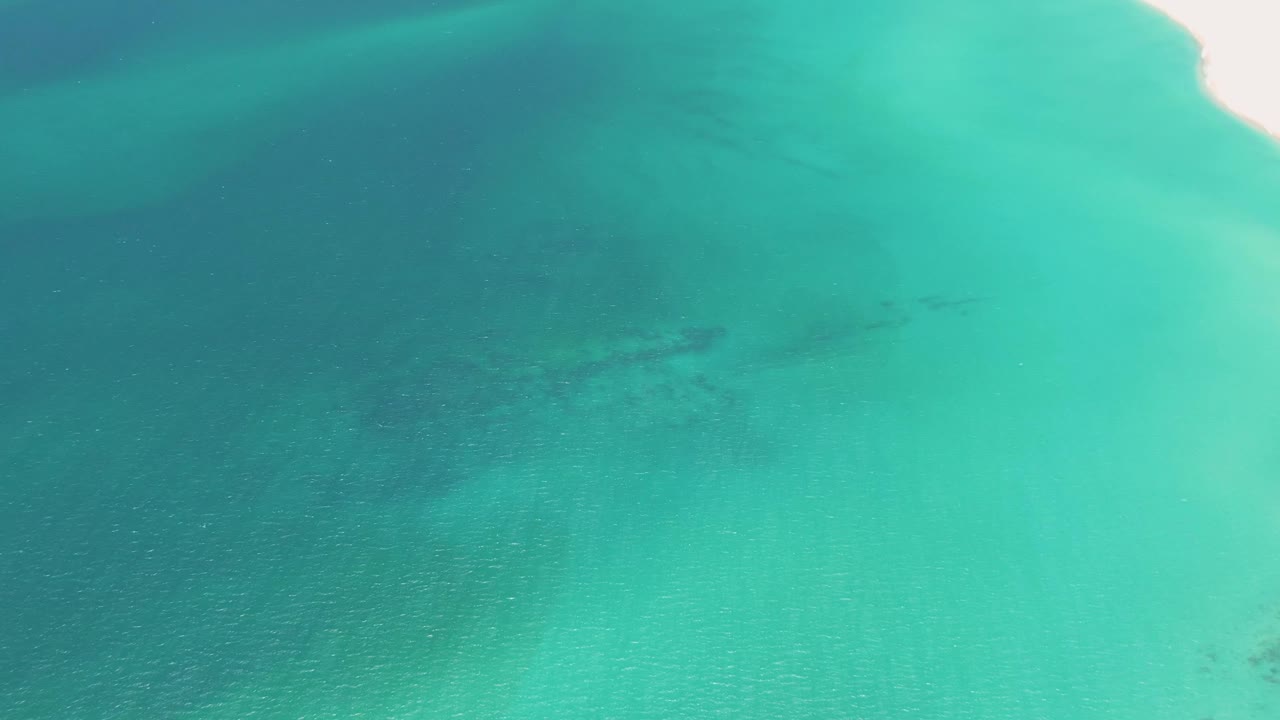 An overhead view at Playa Miramar Los Cabos shows turquoise water and the first bright strip of sandy beach entering at the right edge, with no visible people.
