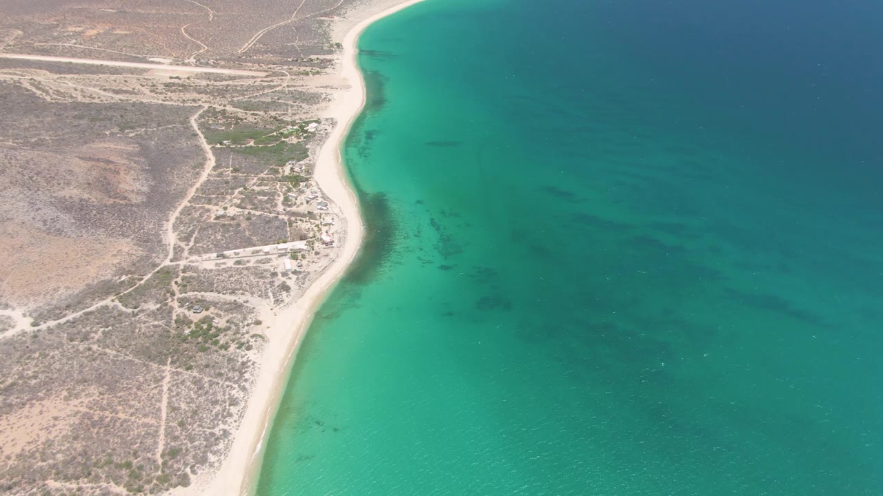 Aerial view at Playa Miramar Los Cabos with the shoreline slightly farther along the coast, pale sand beside turquoise water, dry scrubland, dirt paths, and no visible people.