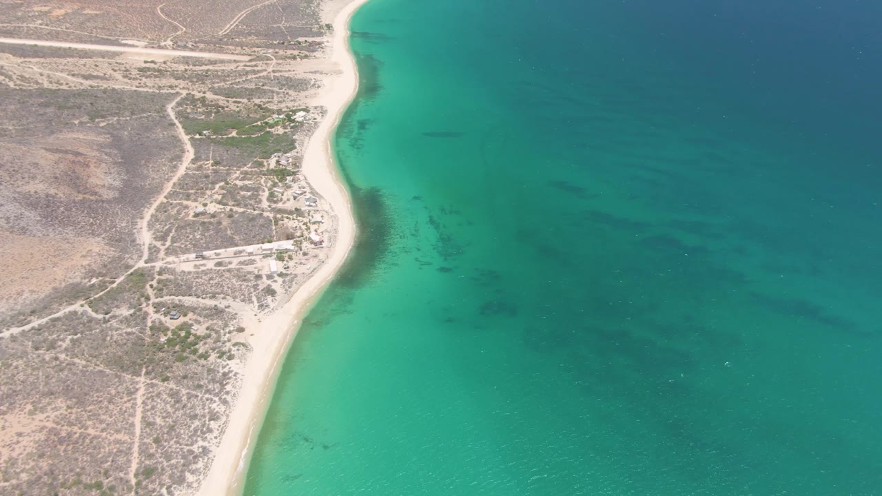 Aerial frame over Playa Miramar Los Cabos where bright turquoise shallows run along the sand, darker water spreads to the right, dry inland terrain lies to the left, and no visible people appear.