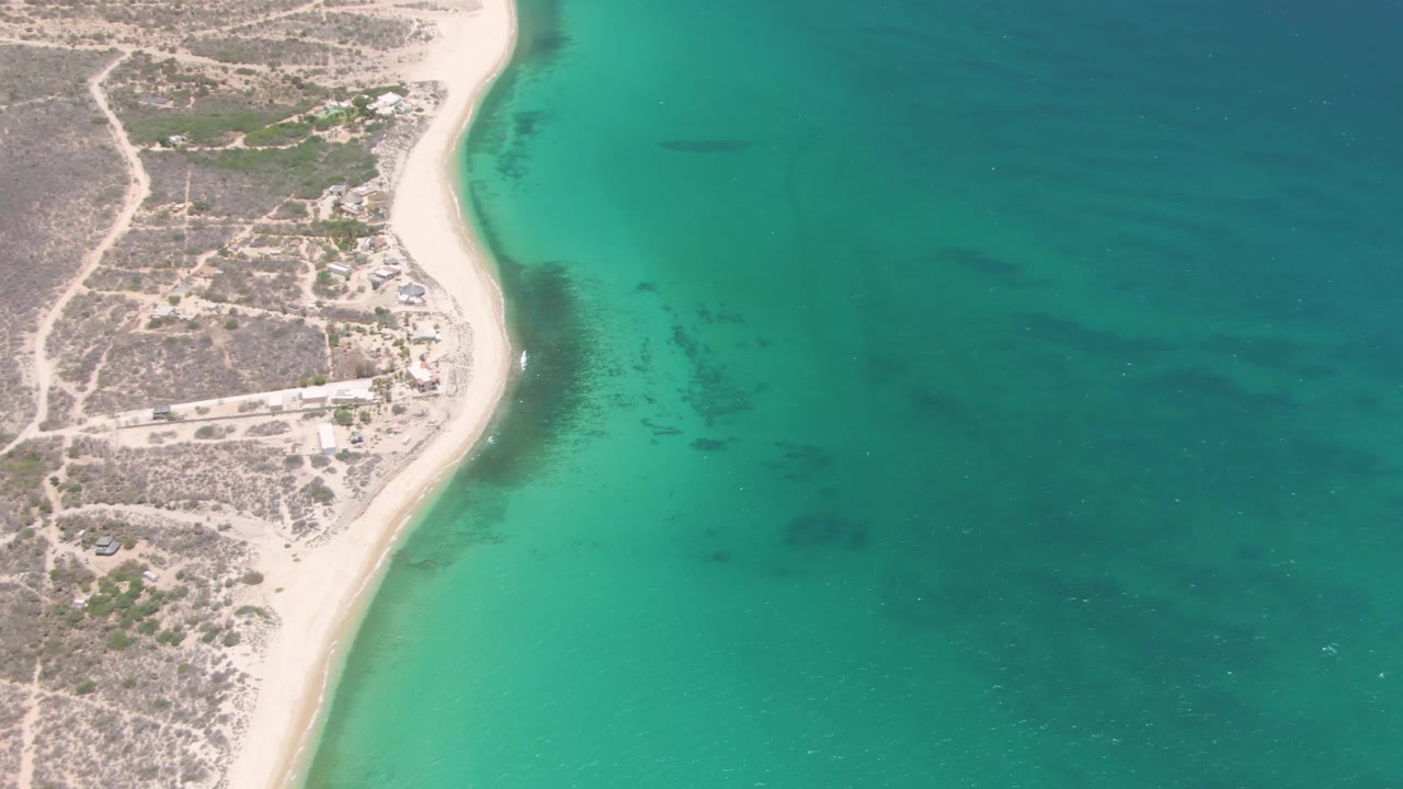 Aerial frame of Playa Miramar, Los Cabos, with pale sand curving along turquoise water, dry coastal land and small structures at left, and no visible people.