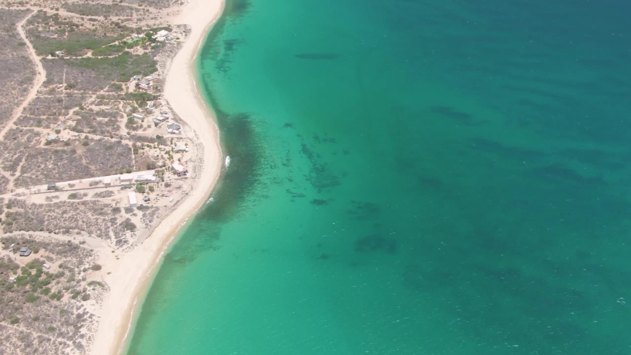 Aerial view over Playa Miramar showing the beach line tighter along the left side, clear turquoise shallows, darker underwater patches, dry land, and no visible people.