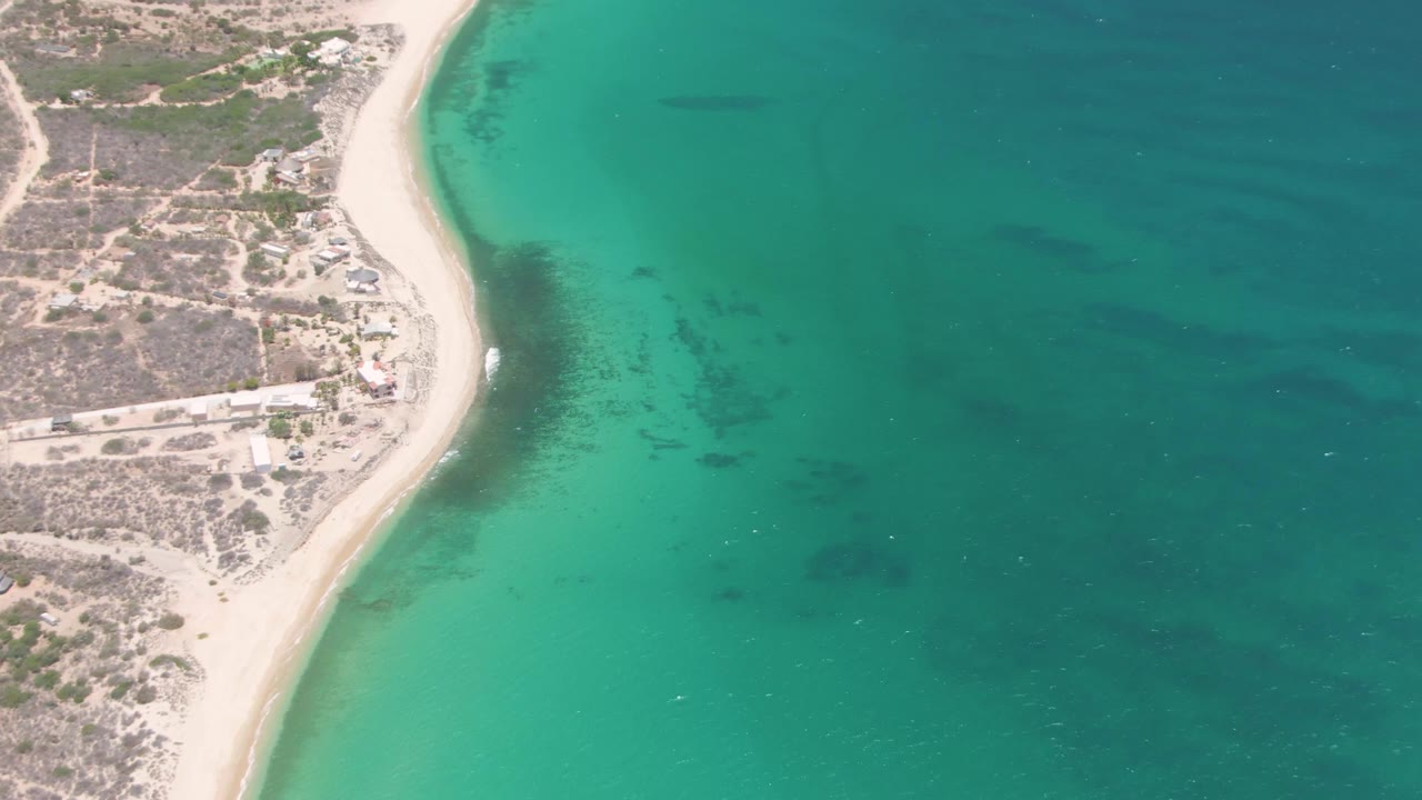 High drone frame of Playa Miramar with the sandy shore running diagonally, small coastal structures and paths on land, broad turquoise water to the right, and no visible people.