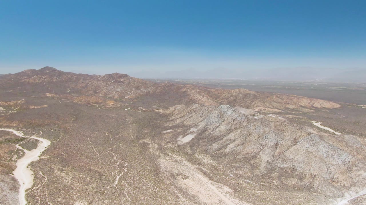 Aerial view of rocky desert ridges and a pale dry riverbed near Playa Miramar Los Cabos, Baja California Sur, with distant mountains and no visible people.