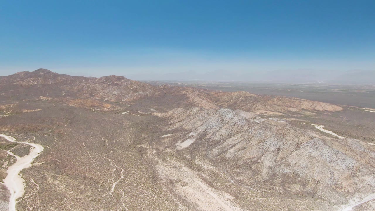Aerial still of a winding dry wash beside rocky ridges in Playa Miramar Los Cabos, with broad blue sky above and no visible people.