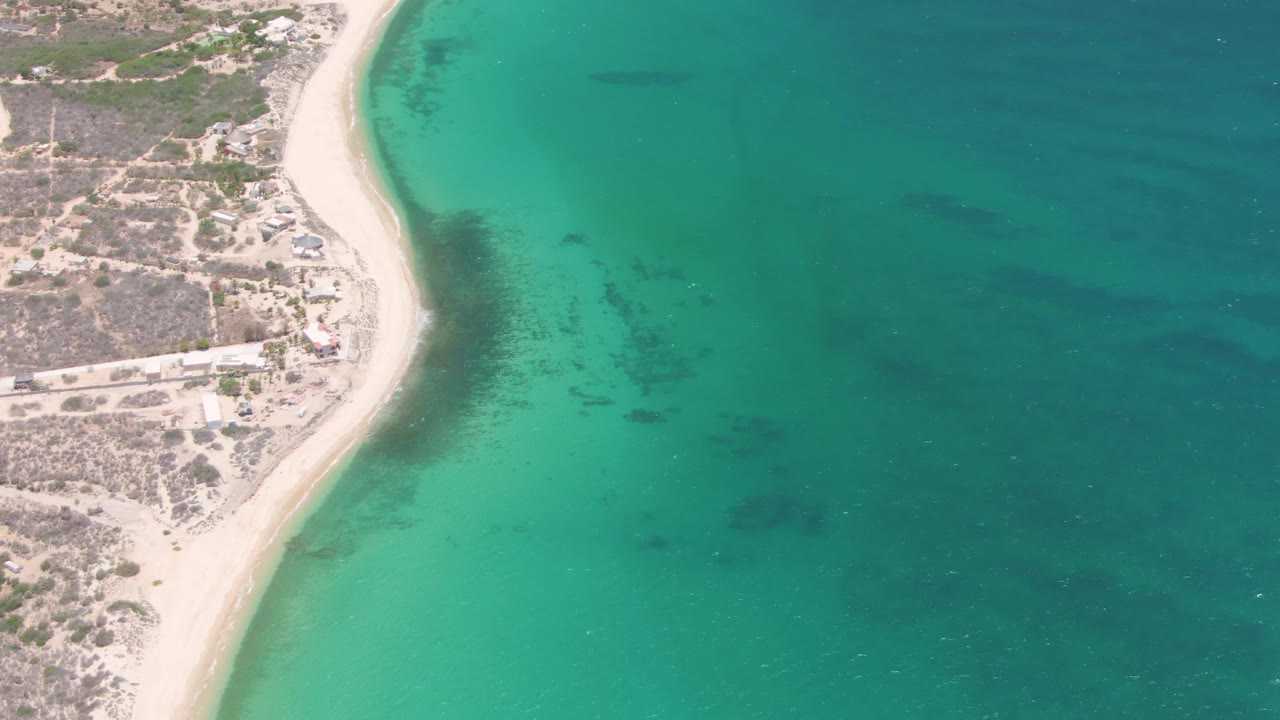 Aerial frame over Playa Miramar showing the beach edge, shallow green-blue water, darker underwater areas, small shoreline structures, and no visible people.