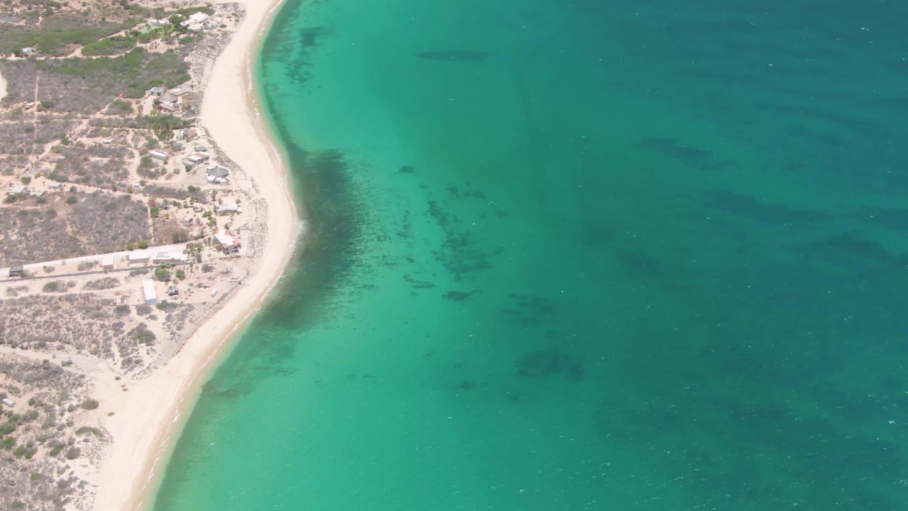 Mid-clip aerial view of Playa Miramar with scattered structures on dry coastal land, a narrow sandy beach, broad turquoise sea, and no visible people.