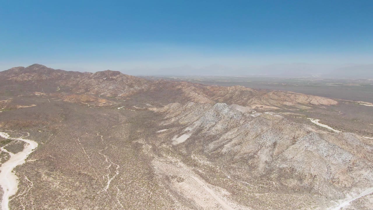 Aerial view over Playa Miramar Los Cabos desert terrain, with a pale dry wash at lower left, rocky ridges across the foreground, distant mountains, and no visible people.