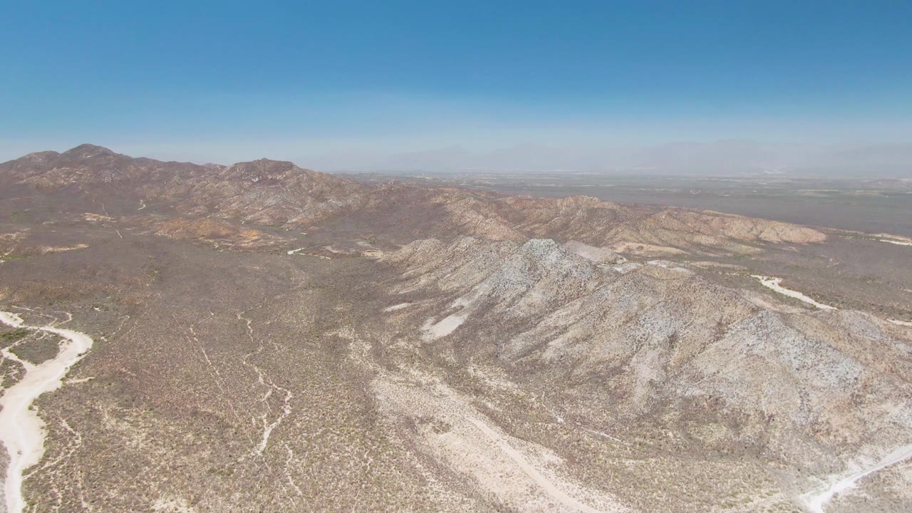 Drone-height view over the Playa Miramar Los Cabos desert, with gray-tan rocky hills in front, branching dry wash channels on the valley floor, and no visible people.