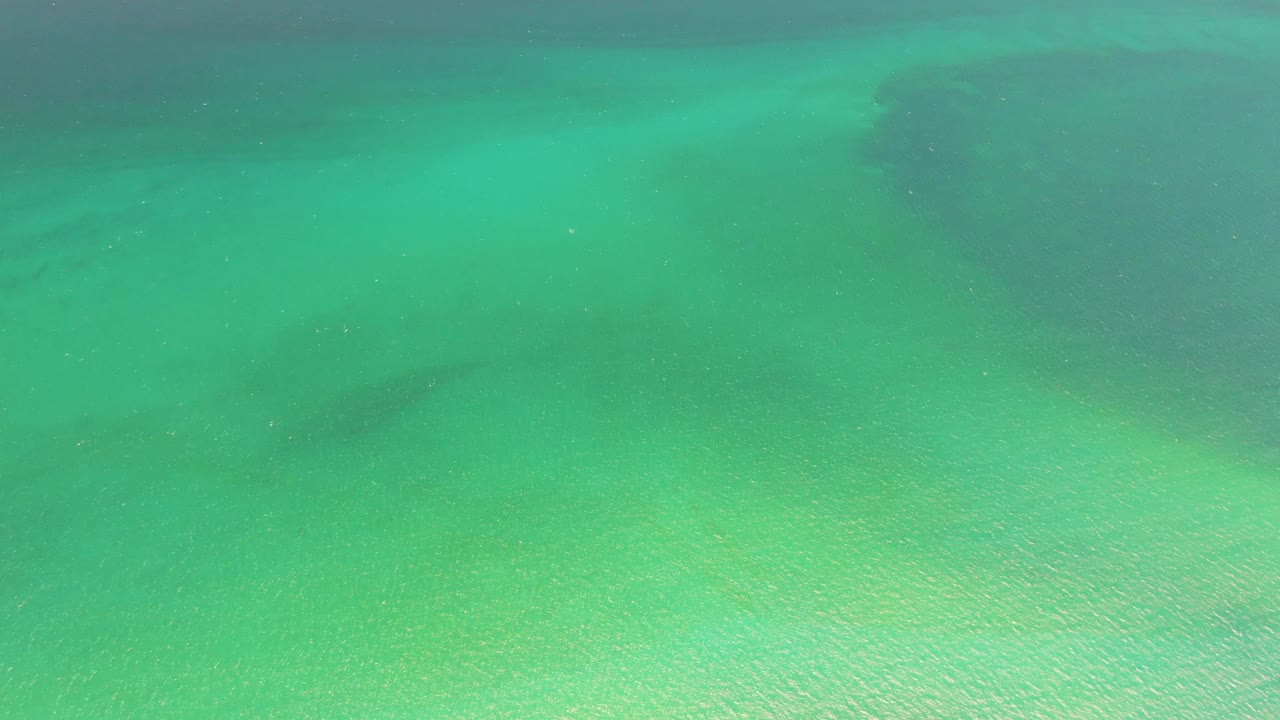 Overhead view of clear turquoise water above the sandy seabed at Playa Miramar Los Cabos, with darker underwater patches near the upper edge and no visible people.