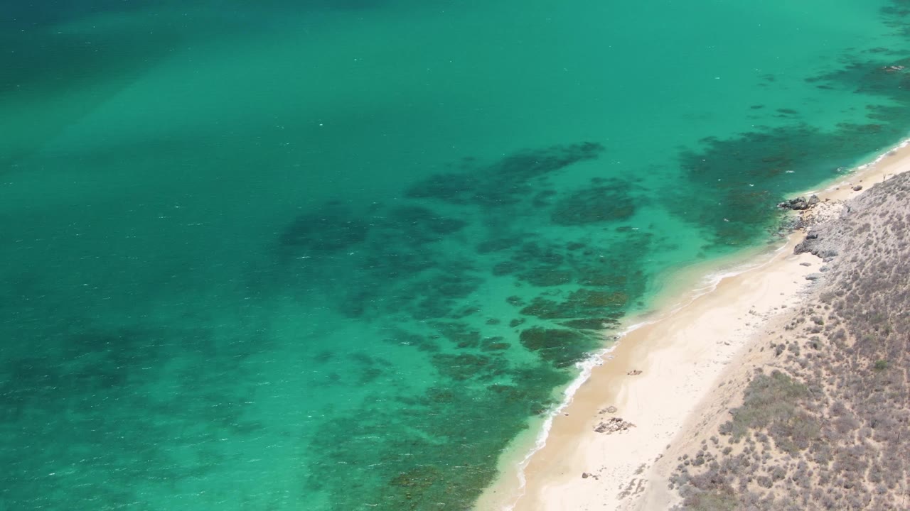 Aerial frame over Playa Miramar Los Cabos showing clear turquoise sea, dark underwater rock or reef patches, pale sand, low surf, and no visible people.
