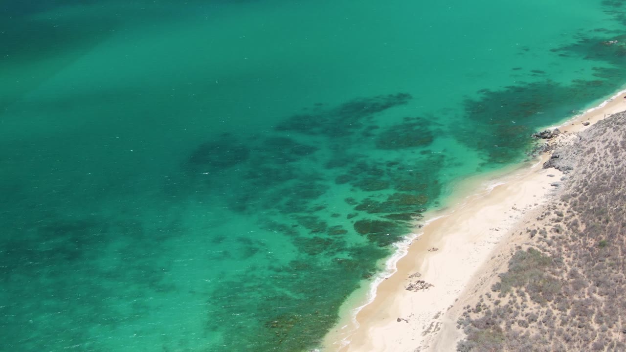 High drone view of the Playa Miramar Los Cabos beach as the shoreline cuts diagonally through turquoise water, dry scrubland, scattered rocks, and no visible people.