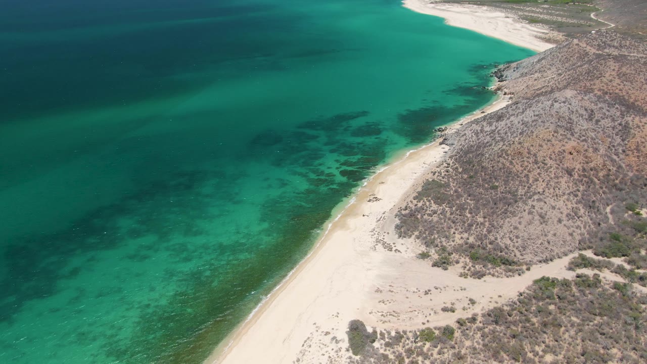 Aerial view over Playa Miramar Los Cabos showing turquoise water, pale sand, rocky hillside vegetation, and no visible people at the opening frame.