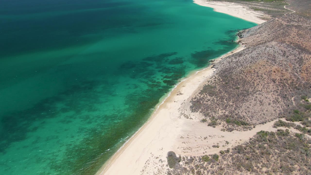 Second frame over Playa Miramar Los Cabos with the surf line slightly lower in frame, clear green-blue water, dry rocky slope, and no visible people.