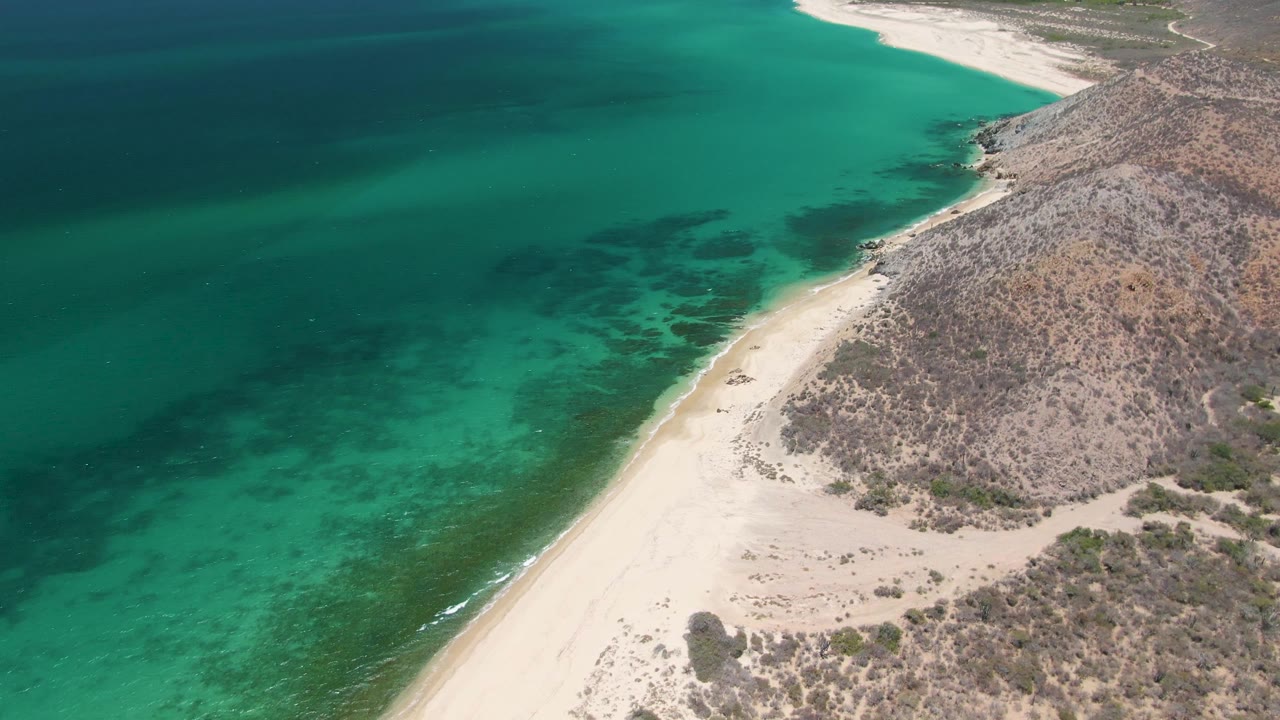 Third frame from Playa Miramar Los Cabos showing more of the curving pale beach at upper right, dark patches under turquoise water, scrubby hillside, and no visible people.