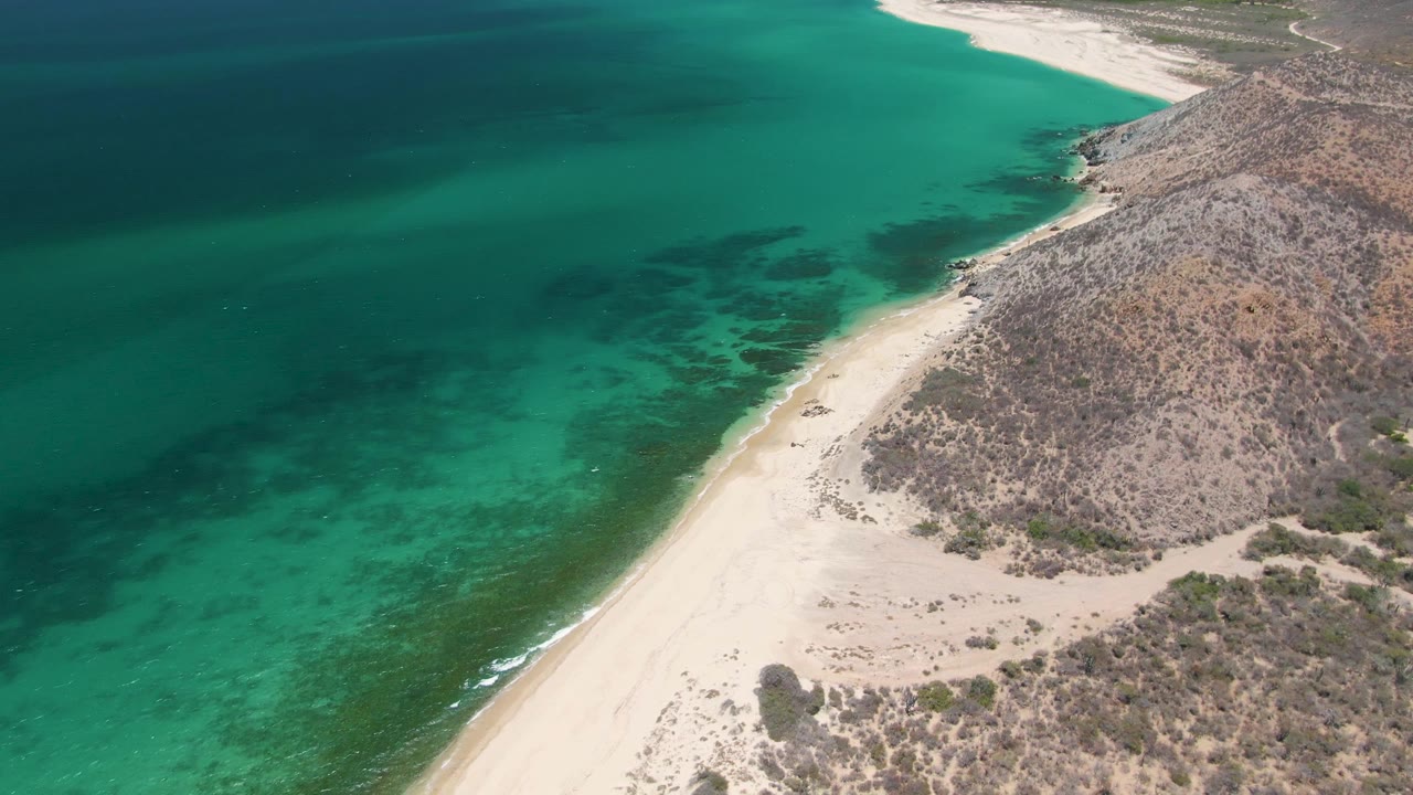 Drone view of empty sandy shore with turquoise water and rocky coastal hillside.