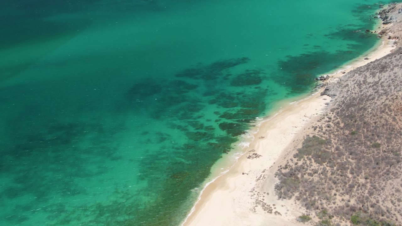 Aerial view of turquoise water beside a sandy beach and rocky hillside.
