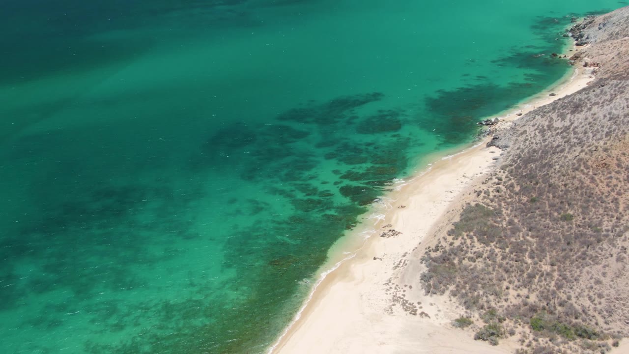 Drone frame over Playa Miramar Los Cabos with clear green-blue water curving along the sandy beach, dry hillside on the right, and no visible people.