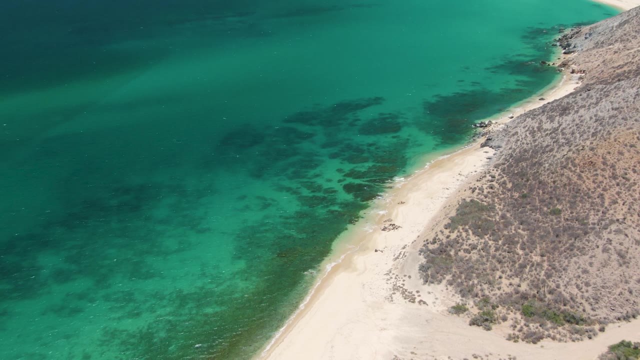 Aerial frame at Playa Miramar Los Cabos showing the beach and brush-covered slope filling more of the right side, with turquoise water and no visible people.