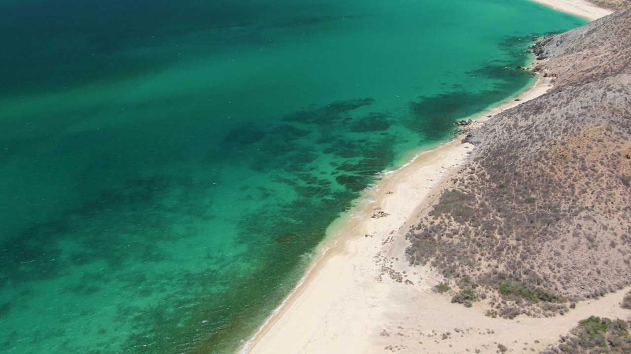 Overhead frame from Playa Miramar Los Cabos showing darker green water offshore, a narrow pale beach strip, rocky dry land, and no visible people.