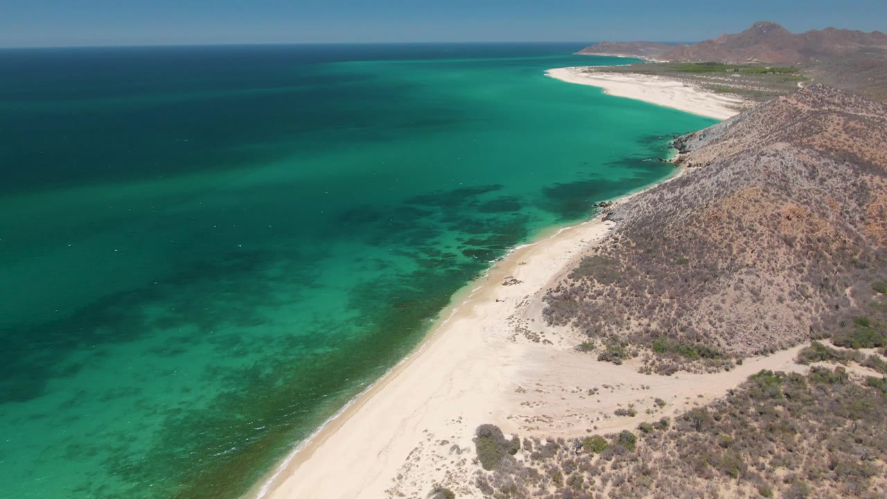 Aerial frame over Playa Miramar Los Cabos showing turquoise shallows beside a white beach, rocky hills on the right, deep blue water offshore, and tiny distant people on the sand.