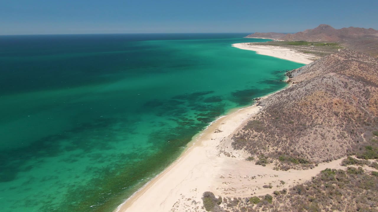 White beach curves along turquoise water with rocky hills and distant mountains.
