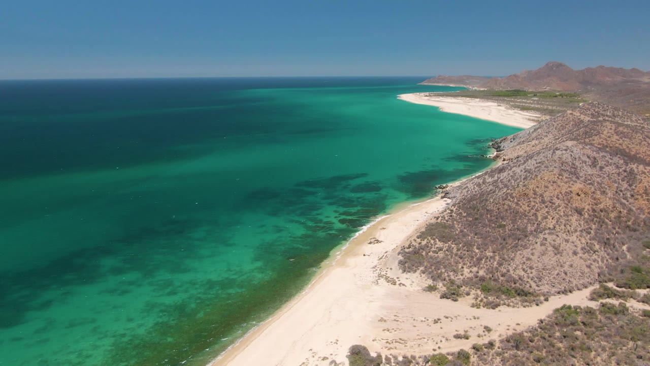 At 0:02, the drone view over Playa Miramar Los Cabos shows more open turquoise water in the foreground, the beach angling along the right side, rocky hills beyond it, and tiny distant people on shore.