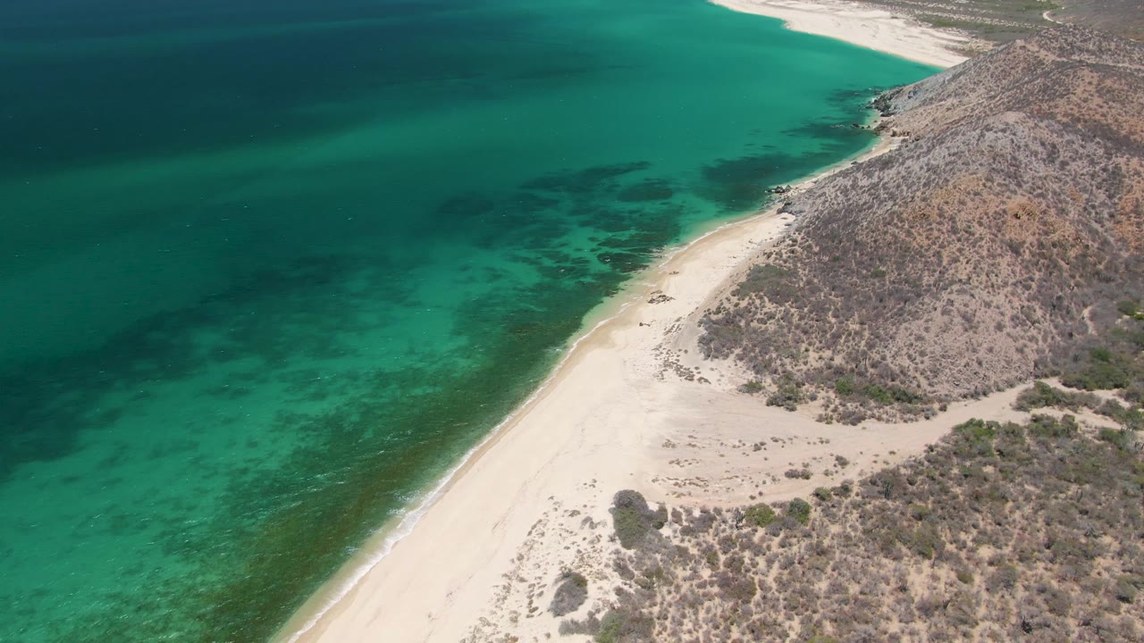 A Playa Miramar Los Cabos aerial frame shows more of the rocky hillside beside the sand while turquoise water and darker submerged patches fill the left side, with no visible people.