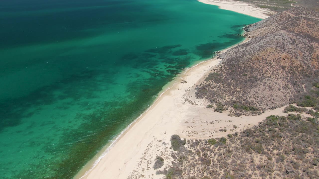 At three seconds, calm turquoise water runs along the sandy Playa Miramar Los Cabos shoreline, with rocky terrain and low brush inland and no visible people.