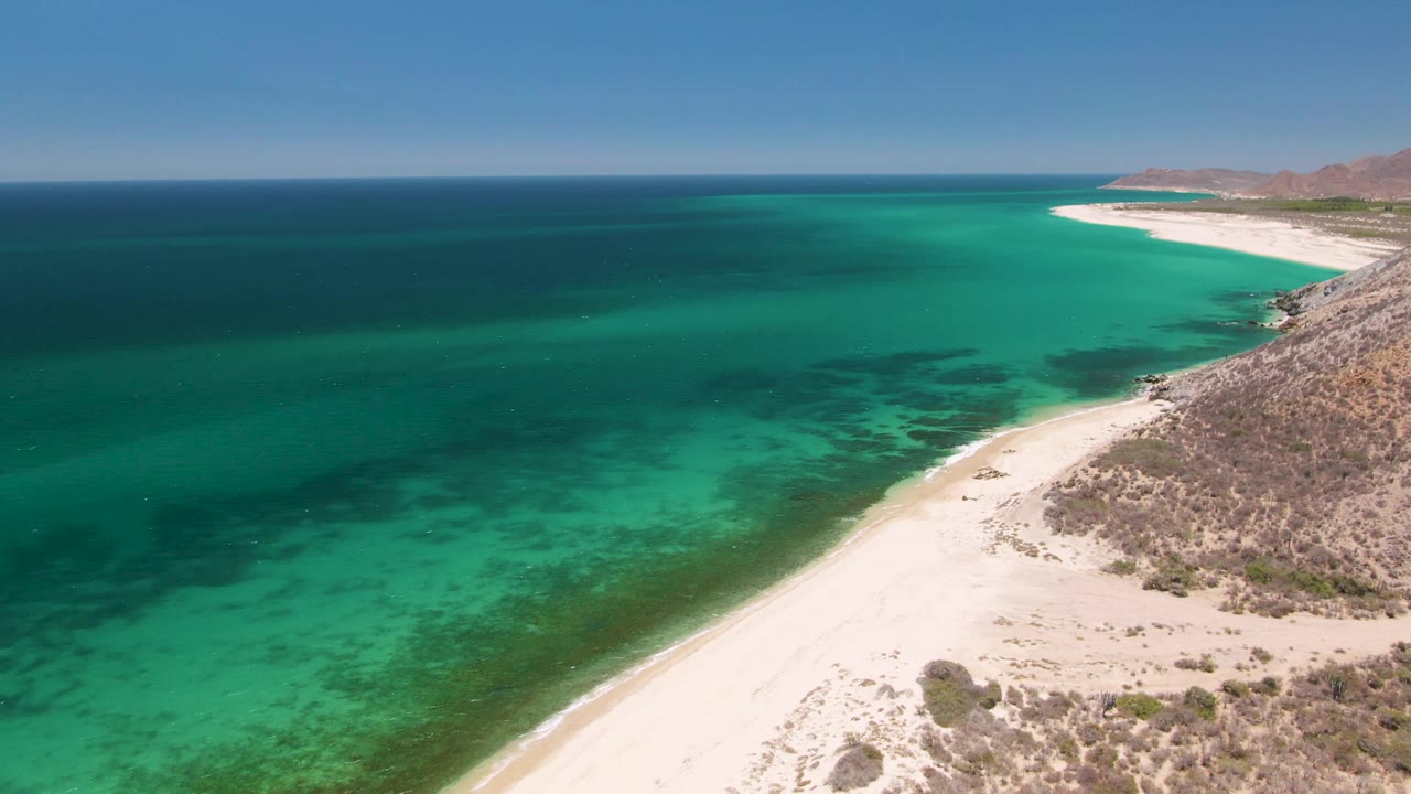 Aerial frame over Playa Miramar Los Cabos shows turquoise water meeting a pale curved beach, dry rocky scrub on the right, distant hills on the horizon, and no visible people.