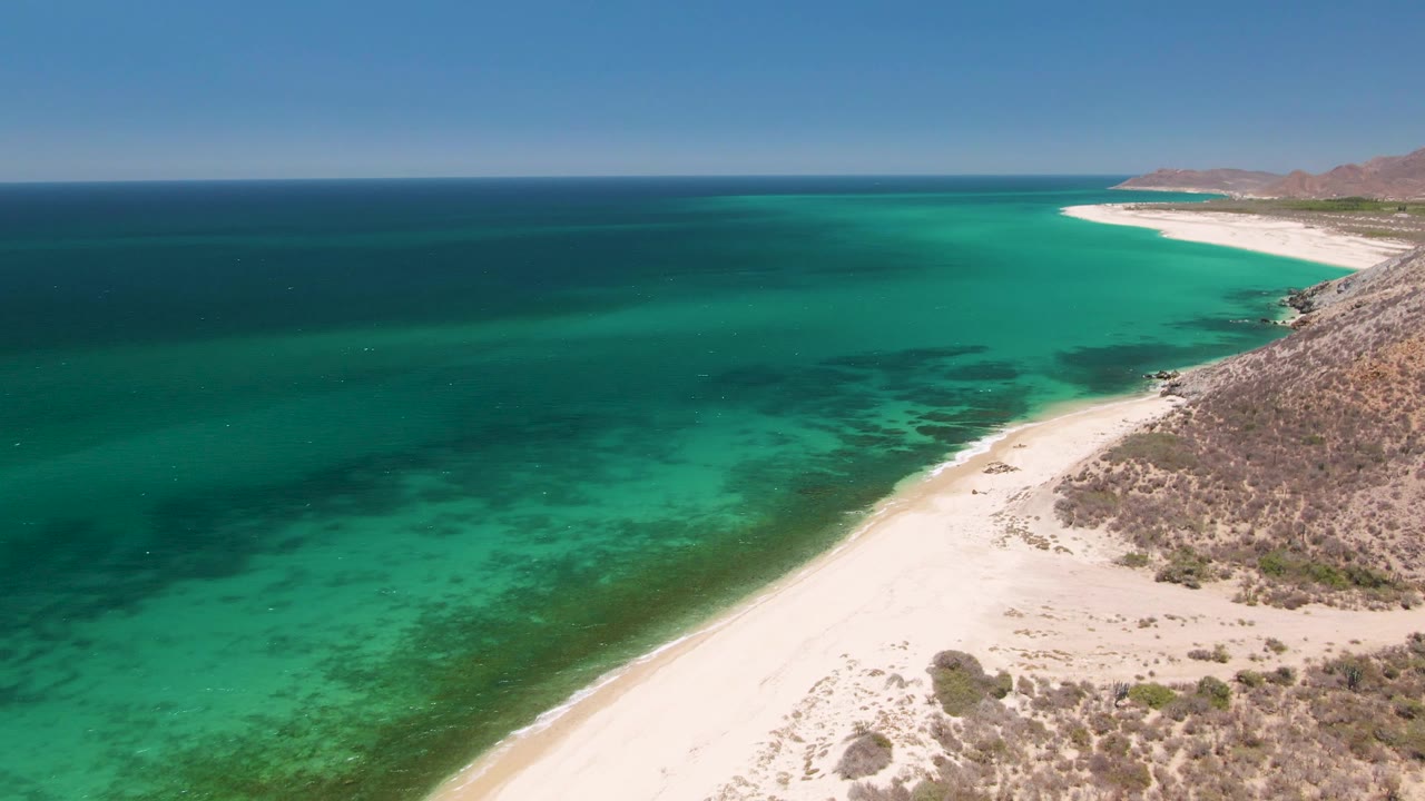 Clear green-blue ocean water runs along a pale beach with rocky desert terrain nearby.