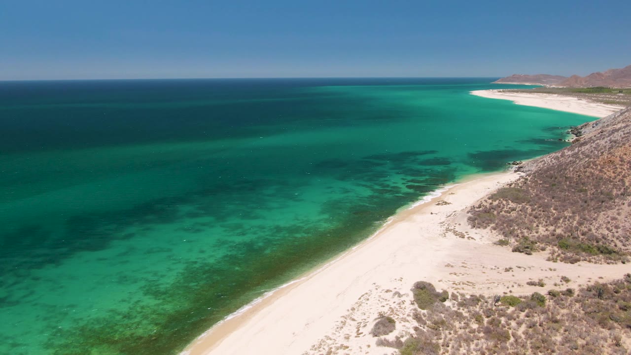 At 0:02, the aerial view over Playa Miramar Los Cabos holds the ocean on the left and the beach on the right, with pale sand foreground, scrub-covered rocky ground, and no visible people.