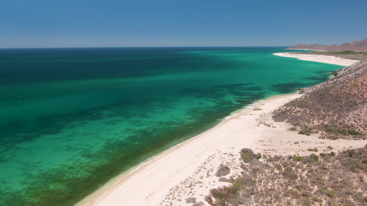 Aerial view of turquoise ocean beside a sandy beach and rocky hillside.