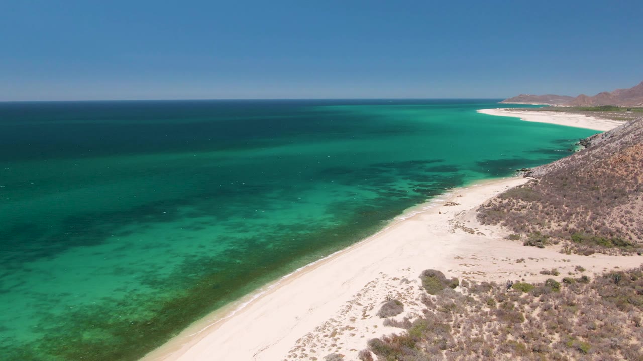 A second aerial frame of Playa Miramar Los Cabos, Mexico, with the shoreline running diagonally below clear turquoise water, rocky hillside at right, distant coast, and no visible people.