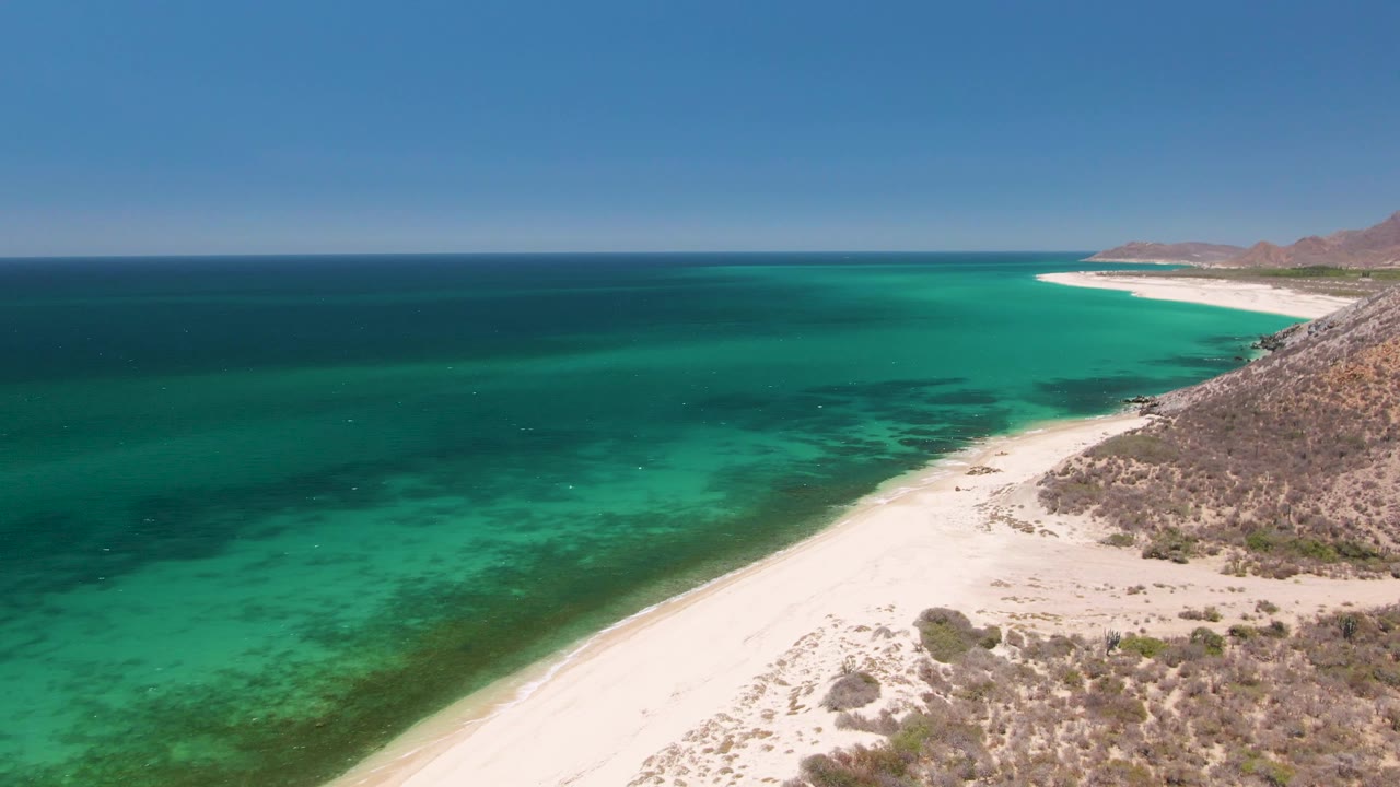 Mid-clip aerial view at Playa Miramar Los Cabos, Mexico, showing more open water across the frame, a curved pale beach at lower right, dry hillside, blue horizon, and no visible people.