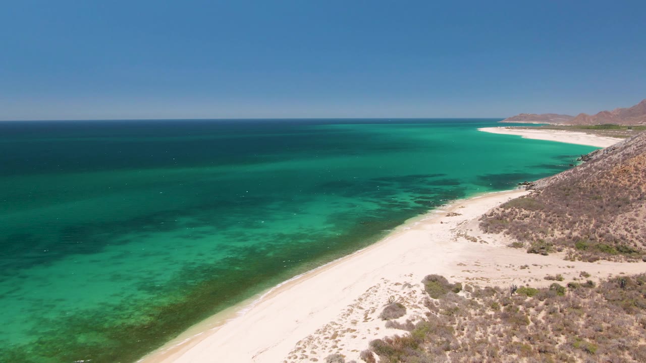 Later aerial frame over Playa Miramar Los Cabos, Mexico, with calm turquoise water on the left, pale sand in the lower frame, scrub-covered rocks on the right, distant coast, and no visible people.