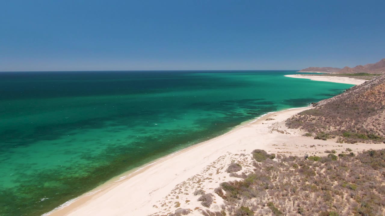 Aerial frame over Playa Miramar Los Cabos showing white sand, turquoise ocean on the left, dry scrubland on the right, low hills in the distance, and no visible people.