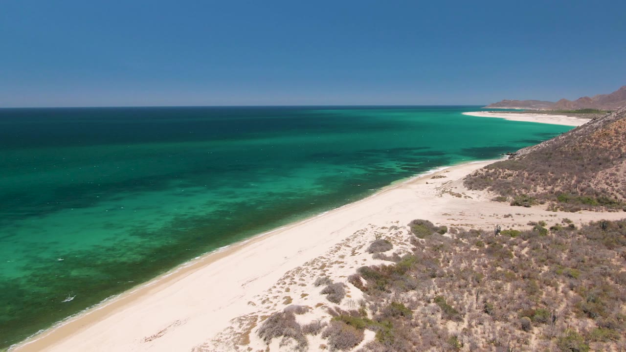 Aerial frame from Playa Miramar Los Cabos showing broad pale sand, textured turquoise water, dry brush and rocky slope along the right side, and no visible people.