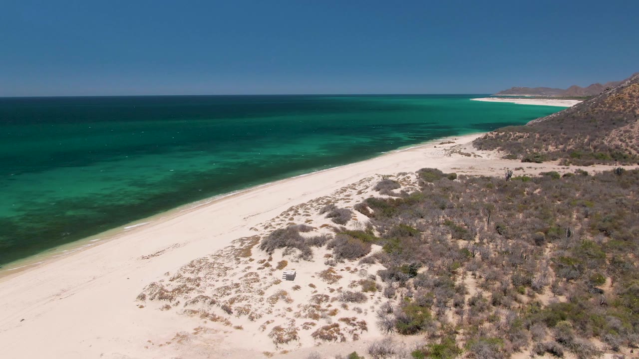 Aerial still from Playa Miramar Los Cabos showing a long white beach, layered turquoise water, dry shrubs, rocky terrain to the right, and no visible people.