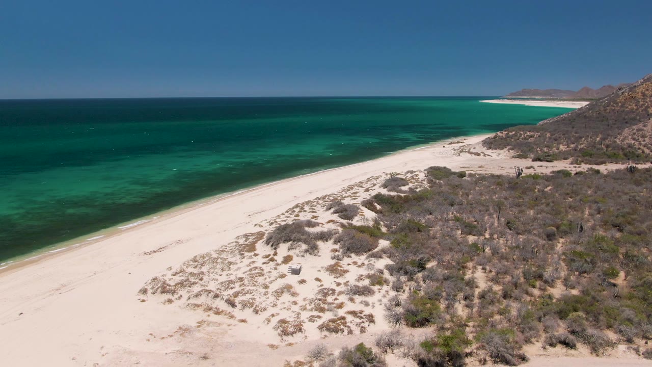 Oblique aerial frame at Playa Miramar Los Cabos with empty white sand in the lower left, turquoise sea beyond it, scrubby dunes in the foreground, and no visible people.