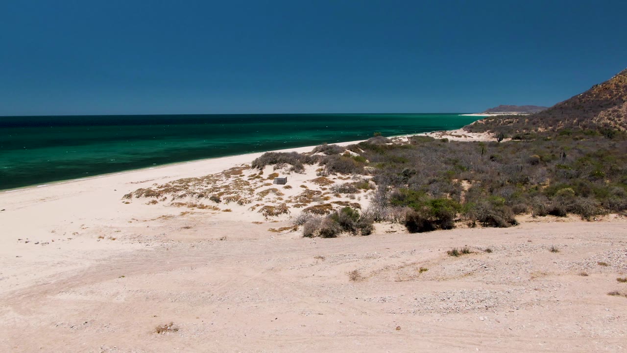 The frame holds an empty Playa Miramar Los Cabos beach with low scrub across the dunes, turquoise ocean at left, distant shoreline beyond, and no visible people.