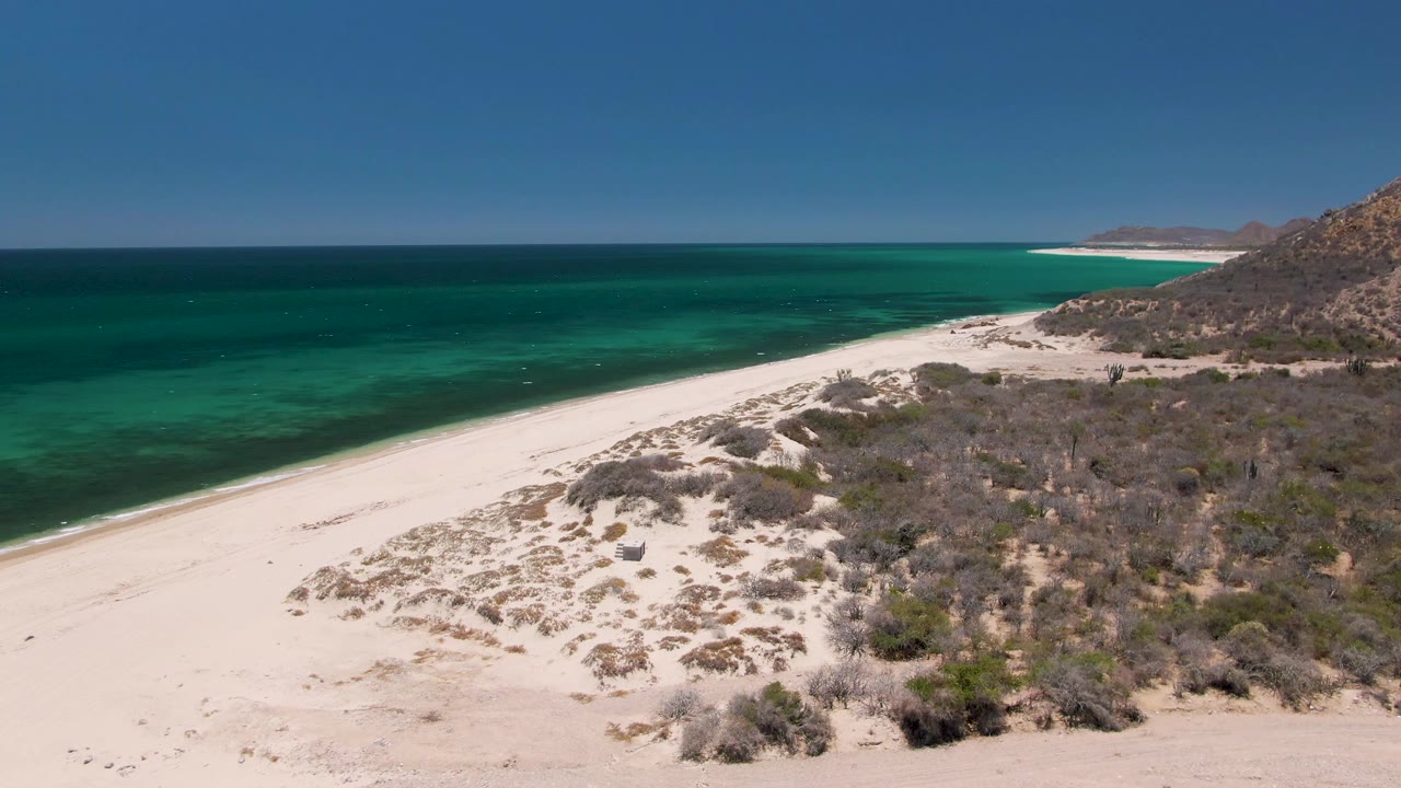 Aerial view of white sand beach, dunes, turquoise ocean, and rocky hillside.
