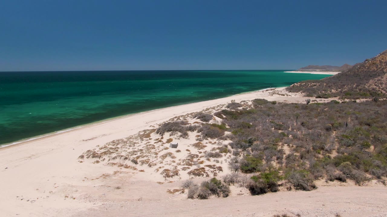 Aerial frame at Playa Miramar Los Cabos showing more of the dune field in front, a clean white shoreline, layered turquoise ocean color, rocky terrain at right, and no visible people.