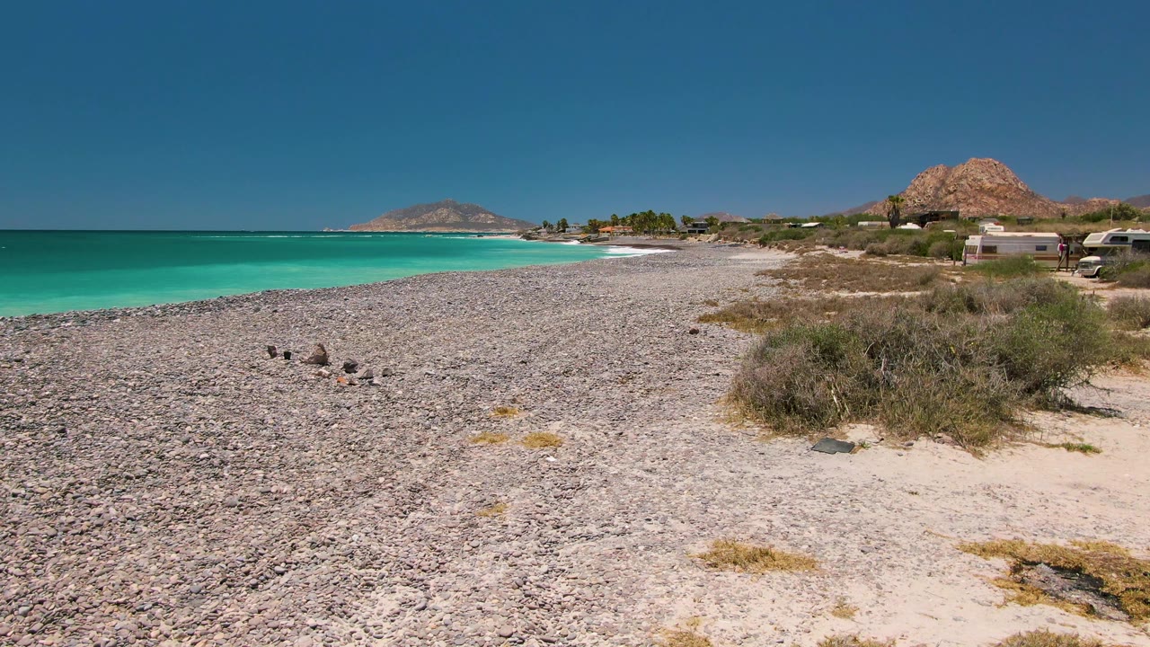 Cabo Pulmo beach frame with turquoise water on the left, pebbled shore in the foreground, dry shrubs and small shoreline structures to the right, birds on the stones, and no visible people.