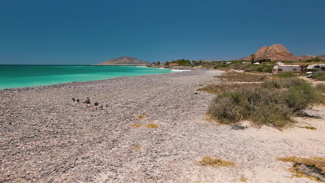 Cabo Pulmo shoreline with the ocean still left of frame, a broad pebbled beach through the center, dry vegetation and low structures to the right, birds near the foreground stones, and no visible people.