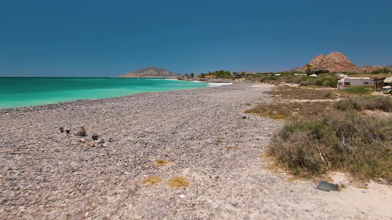 Cabo Pulmo coastal frame where the camera has shifted slightly down the pebbled shore, with turquoise water at left, dry scrub at right, small structures beyond the beach, distant hills, and no visible people.