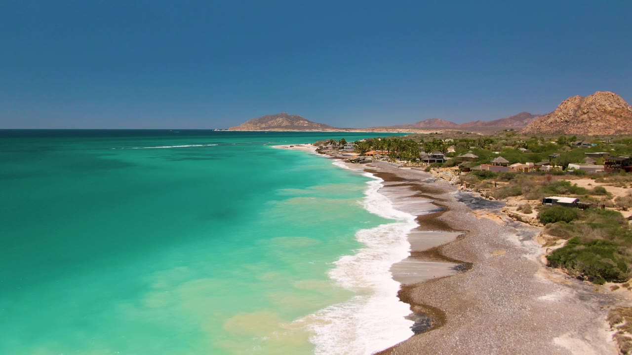 Aerial view in Cabo Pulmo, Baja California Sur, with turquoise water on the left, white surf reaching a sandy and pebbly beach, small structures near sparse vegetation, rocky hills in back, and no visible people.